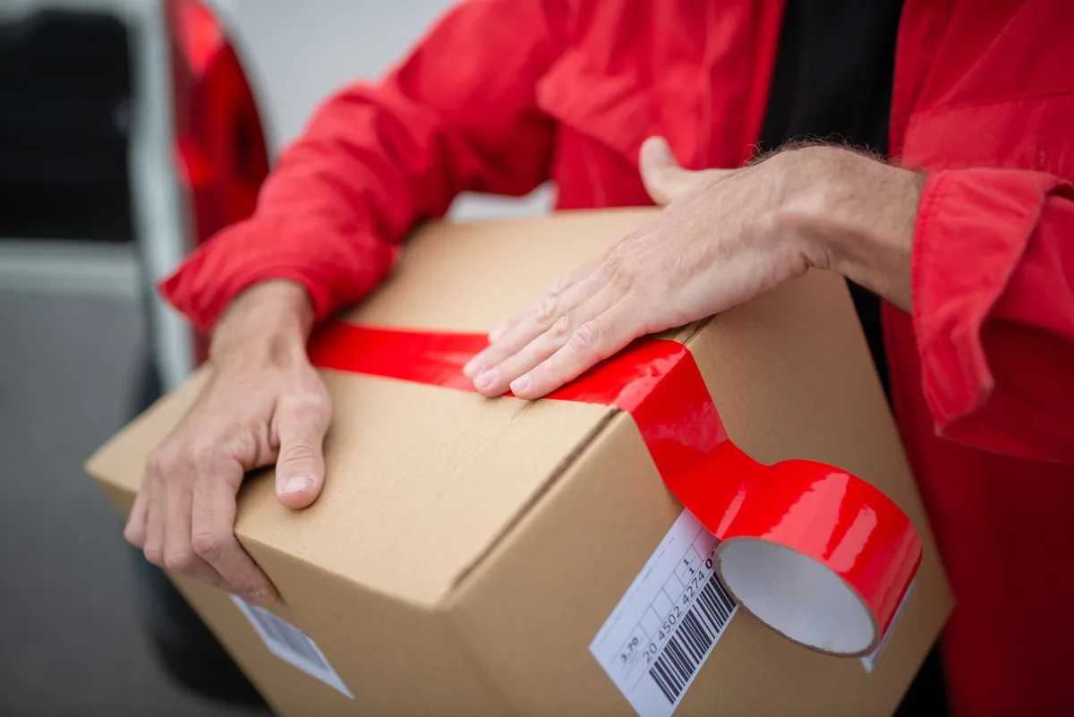 Tangan menutup paket dengan lakban merah, label resi dan barcode terlihat menempel di kotak