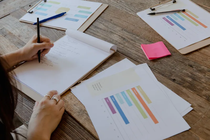 Tangan menulis catatan perhitungan margin dengan grafik warna-warni di atas meja kerja