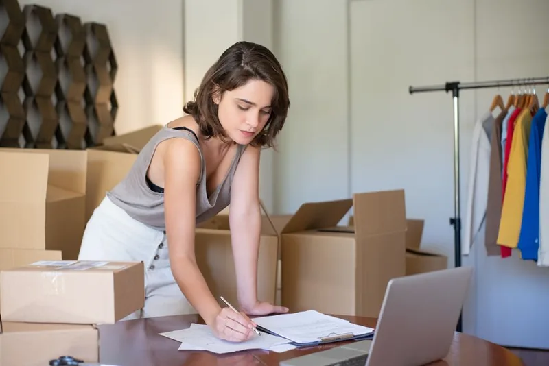 Penjual perempuan mencatat pesanan di meja kerja dikelilingi kardus paket, rak baju, dan laptop