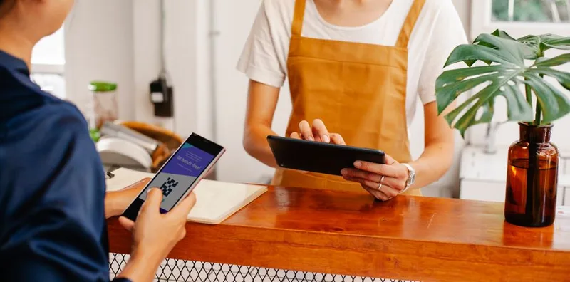 Pelanggan menunjukkan kode QR di layar ponsel kepada kasir yang memegang tablet di konter kafe